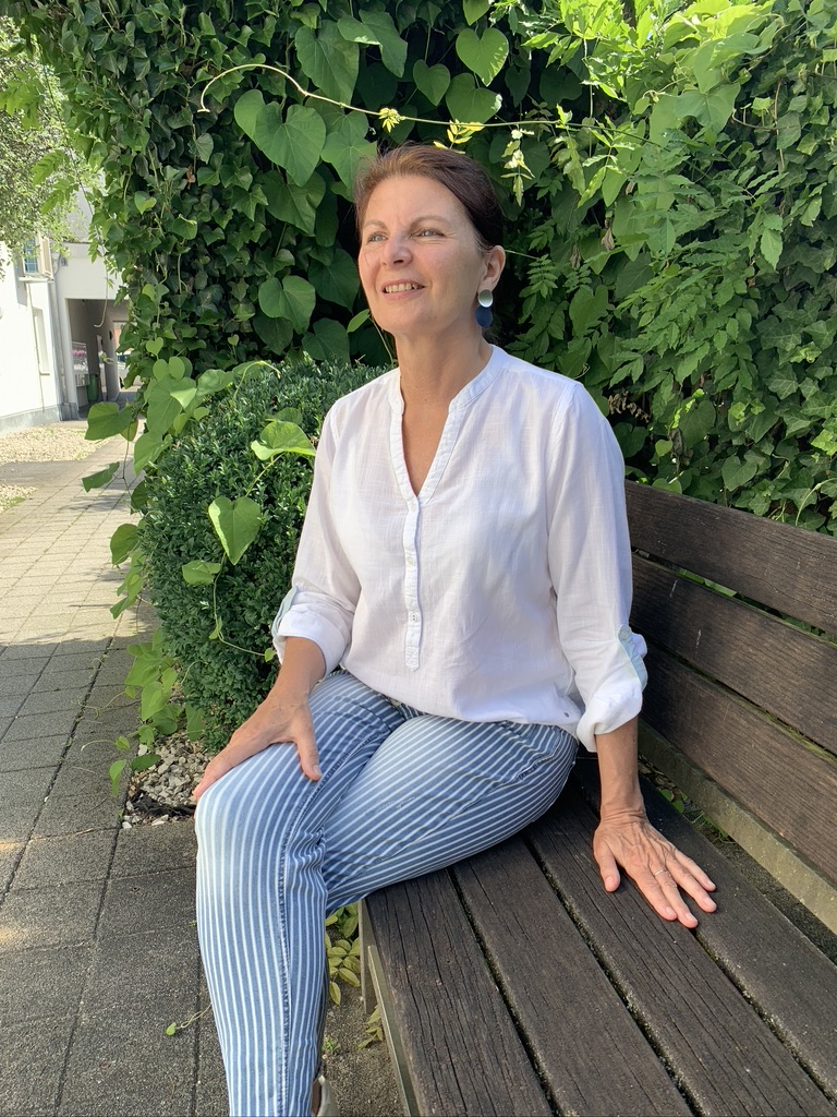 Heilpraktikerin Barbara Schütz sitzt lächelnd auf einer Holzbank vor einer Efeuwand