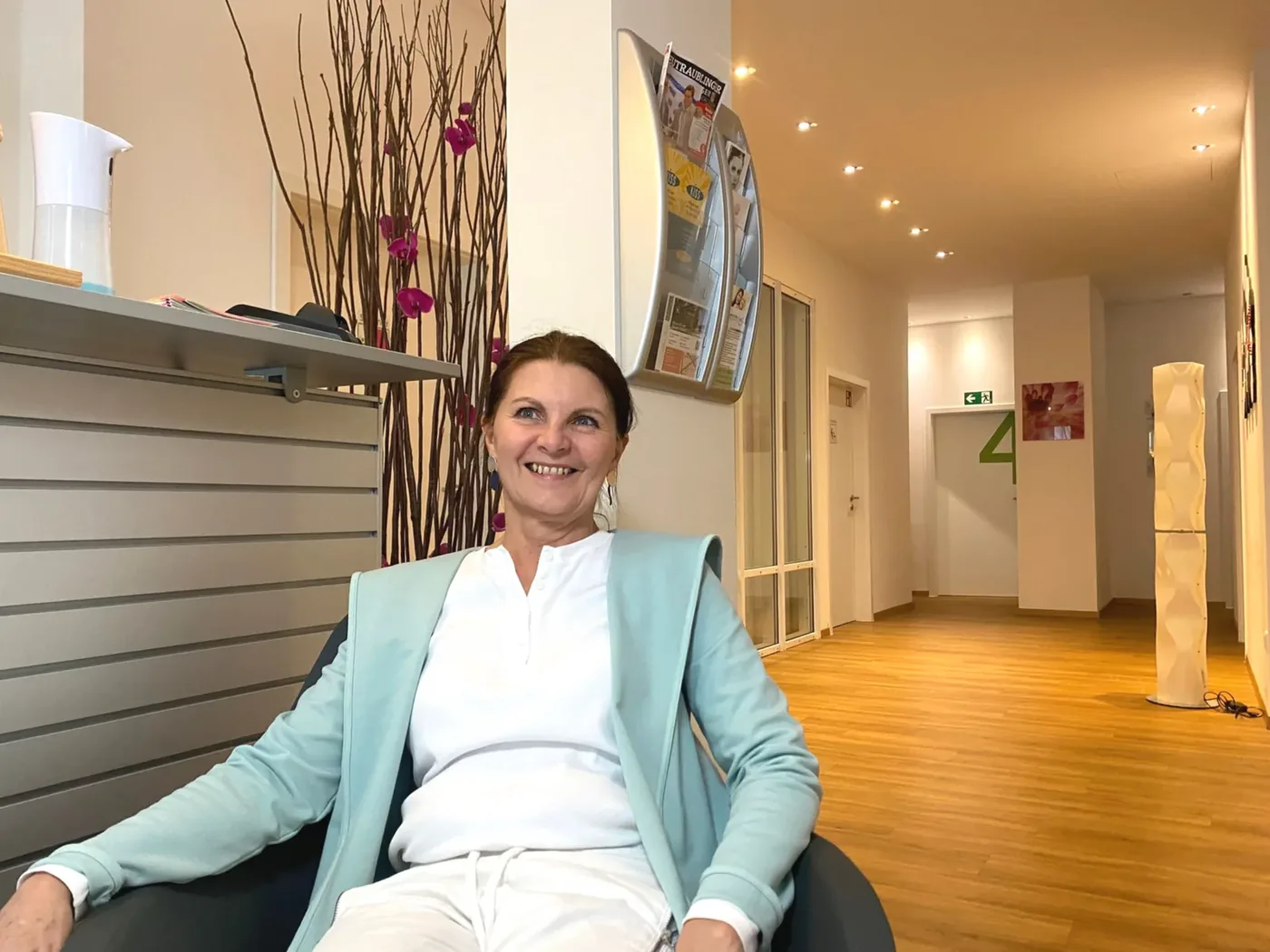 Heilpraktikerin Barbara Schütz sitzt im Praxisflur, im Hintergrund Empfangstresen und Wartebereich